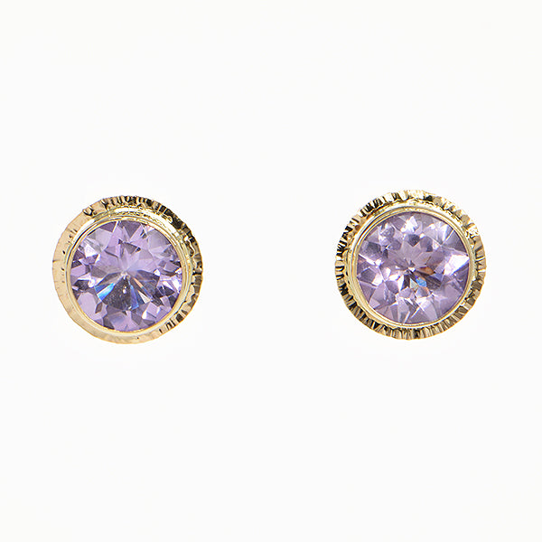 A pair of gold round stud earrings with faceted light purple gemstones, set in a textured gold bezel on a plain white background.