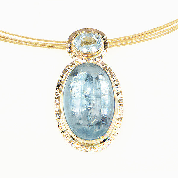 A close-up of a gold necklace pendant featuring a large oval light blue gemstone and a smaller oval light blue gemstone, set on a white background.
