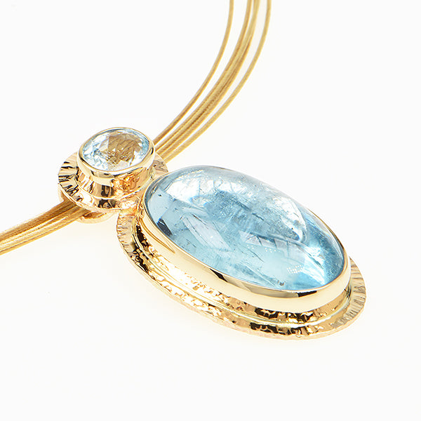 A close-up of a gold necklace pendant featuring a large oval light blue gemstone and a smaller oval light blue gemstone, set on a white background.
