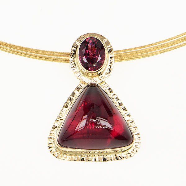 A gold pendant necklace featuring a large triangular deep red gemstone and a smaller oval matching red gemstone, shown on a clean white background.