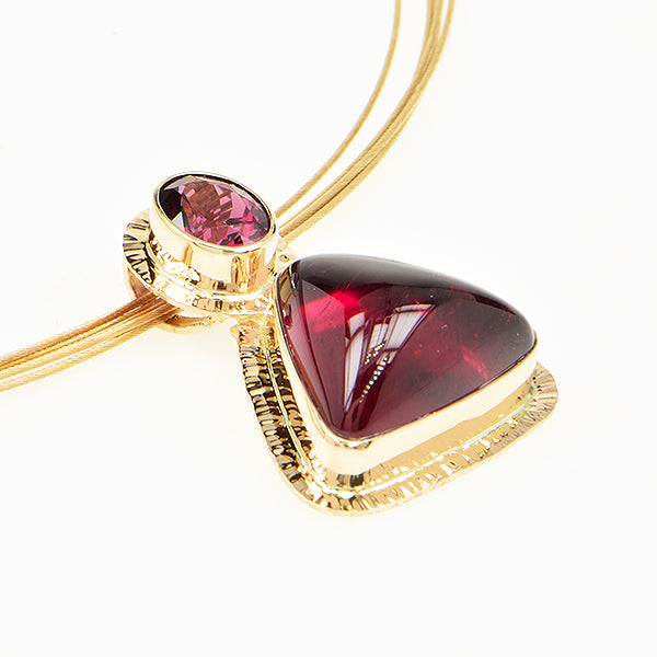 A gold pendant necklace featuring a large triangular deep red gemstone and a smaller oval matching red gemstone, shown on a clean white background.